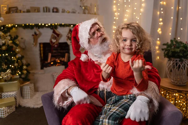 Funny Santa Claus sentado en su rockero con un niño lindo sentado en su rodilla. Concepto de espíritu navideño, fiestas y celebraciones Imagen De Stock