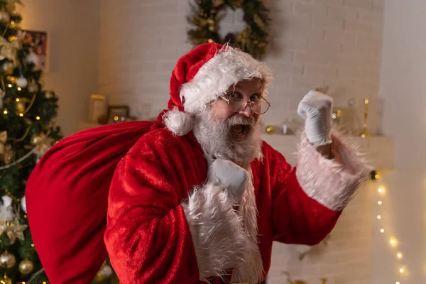 Tradycyjny Święty Mikołaj trzyma torbę z prezentami w domu w pobliżu choinki i kominka. Duch Świąt, magia, koncepcja marzeń — Zdjęcie stockowe