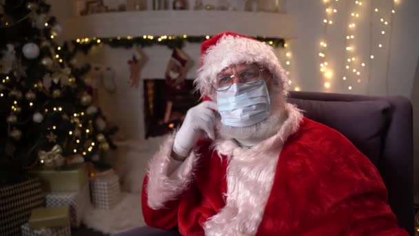 Babbo Natale divertente indossa una maschera medica protettiva mentre seduto su una sedia vicino al camino e all'albero di Natale. Covid 19, protezione contro il coronavirus. — Video Stock