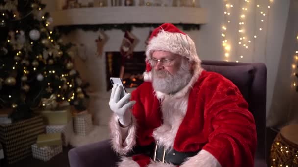 Babbo Natale è seduto su una sedia davanti al camino di notte e utilizzando il telefono. Natale come concetto di vacanza. — Video Stock