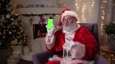 Mutlu Noel Baba, Noel ağacı ve şöminenin yanındaki sandalyede oturuyor ve yeşil ekranlı cep telefonunu gösteriyor. Noel ruhu, tatiller ve kutlamalar konsepti 4K görüntüler
