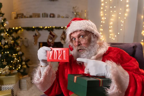 Portret zabawnego Mikołaja w okularach, siedzącego w bujanym fotelu obok choinki i trzymającego tabliczkę czekolady z napisem 2021. Koncepcja ducha Bożego Narodzenia, święta i — Zdjęcie stockowe