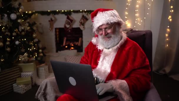 Der moderne Weihnachtsmann. Fröhlicher Weihnachtsmann am Laptop und lächelnd auf seinem Stuhl sitzend mit Kamin und Weihnachtsbaum im Hintergrund. — Stockvideo