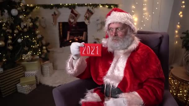 Portret zabawnego Mikołaja w okularach, siedzącego w bujanym fotelu obok choinki i trzymającego tabliczkę czekolady z napisem 2021. Koncepcja ducha Bożego Narodzenia, święta i — Wideo stockowe