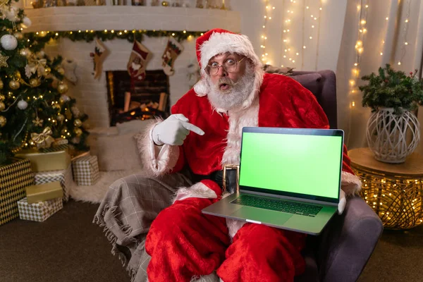 Śmieszny Święty Mikołaj w okularach siedzący w rockerze koło choinki. posiada laptopa z zielonym ekranem i przyciąga uwagę na to. Świąteczny nastrój, święta i uroczystości koncepcja 4k materiał — Zdjęcie stockowe