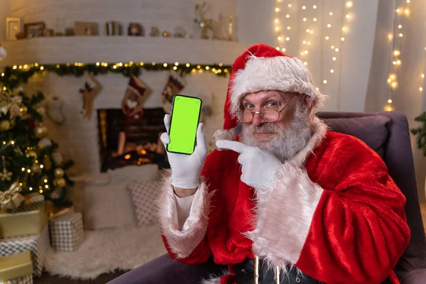 Feliz Santa Claus sentado en la silla cerca del árbol de Navidad y la chimenea, mostrando el teléfono móvil con pantalla verde. Concepto de espíritu navideño, fiestas y celebraciones Fotos De Stock