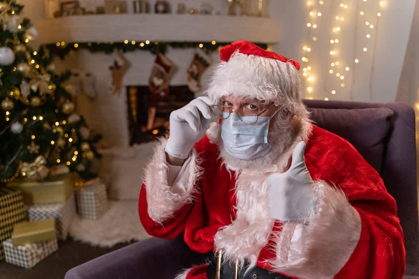 Santa Claus en una máscara médica protectora mientras está sentado en una silla junto a la chimenea y el árbol de Navidad. Covid 19, protección contra el coronavirus. Imagen De Stock