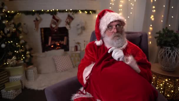 Funny Santa Claus siting on a chair near the Christmas tree unties the bag and jokes takes out a gift — Stock Video