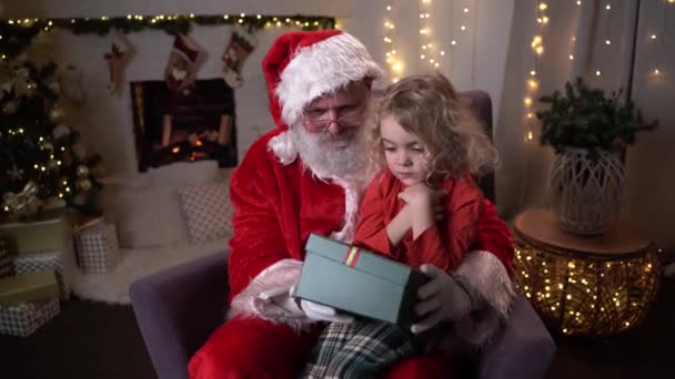 Lustiger Weihnachtsmann sitzt auf seiner Wippe, kleiner süßer Junge sitzt auf seinem Knie und öffnet ein Geschenk mit etwas Besonderem zusammen. Weihnachtsstimmung, Feiertage und Feste — Stockvideo