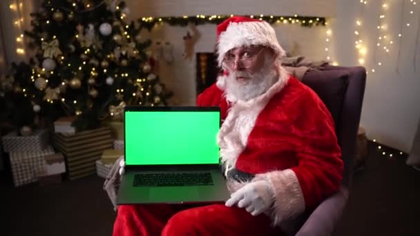 Père Noël dans des lunettes assis dans son rocker près de l'arbre de Noël. tient un ordinateur portable avec écran vert et attire l'attention sur elle. Esprit de Noël, vacances et célébrations concept 4k images — Video