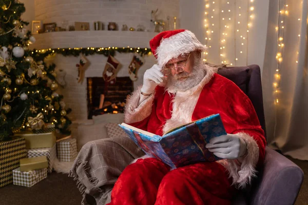 Śmieszna klauzula Mikołaja siedzi obok kominka i choinki czytając magiczną książkę ze świecącymi kartkami - duch Bożego Narodzenia, magia, koncepcja marzeń — Zdjęcie stockowe