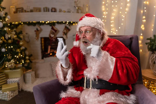 Święty Mikołaj siedzi w nocy na krześle przed kominkiem i używa telefonu. Boże Narodzenie jako koncepcja święta. — Zdjęcie stockowe