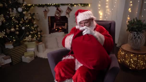 Funny Santa Claus sidder på en stol i nærheden af juletræet løsner posen og vittigheder tager en gave – Stock-video
