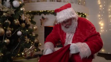 Noel Baba kırmızı bir çantadan hediyeleri alır ve arka planda şömine olan Noel ağacının altına koyar. Noel ruhu, sihir, rüya konsepti 4K görüntüler