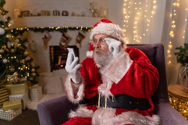 Santa Claus está sentado en una silla frente a la chimenea por la noche y usando el teléfono. Navidad como concepto de vacaciones. — Foto de Stock