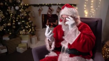 Mutlu Noel Baba, Noel ağacı ve şöminenin yanındaki sandalyede oturuyor ve yeşil ekranlı cep telefonunu gösteriyor. Noel ruhu, tatiller ve kutlamalar konsepti 4K görüntüler