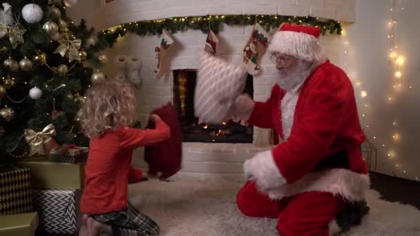 Julemanden og en lille dreng jokingly kæmpe med puder og grine ved pejsen ved siden af juletræet. Julestemning, magi, drømmekoncept 4k optagelser – Stock-video