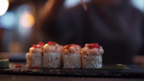 Суши на фоне ресторанных огней. Разнообразие видов суши с красной икрой, рыбой, филадельфийским сыром и палочками крупным планом. Набор вкусных японских суши-роллов на каменной доске — стоковое видео