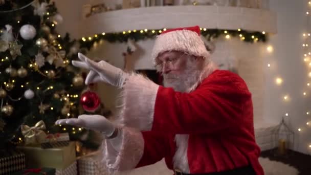 Funny Santa Claus bromea con juguetes de bolas de Navidad al lado del árbol de Navidad en el fondo de la chimenea — Vídeo de stock
