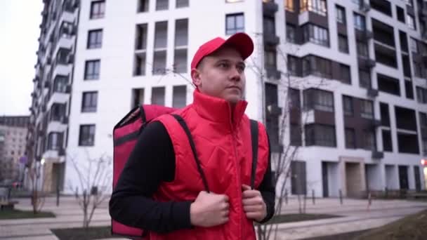Dostawca w czerwonym mundurze przechadzający się nowoczesnymi budynkami ulicą z torbą termiczną. Zbliżenie Średni strzał — Wideo stockowe