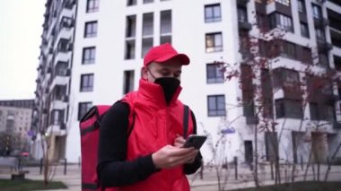 Modern binalarda yürürken koruyucu maske takan ve kırmızı üniformalı bir kurye. Telefonundaki müşteri adresini kontrol ediyor. Virüs salgını sırasında yemek dağıtımı yapıyor. Coronavirus, COVID-19, güvenli teslimat