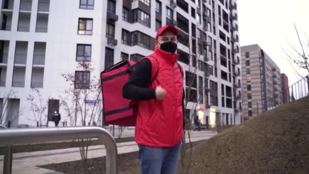 Retrato de repartidor tipo con uniforme rojo y máscara protectora con bolsa térmica de los edificios modernos de fondo. Coronavirus, COVID-19, entrega segura — Vídeos de Stock