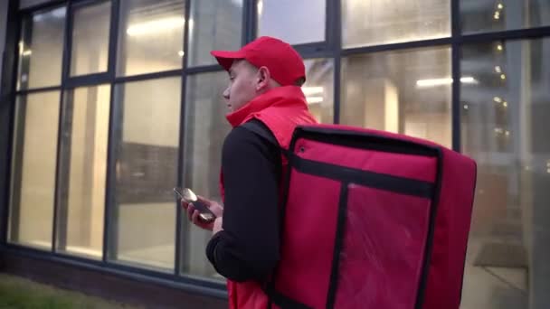 Dostawca w czerwonym mundurze przechadzający się nowoczesnymi budynkami ulicą z torbą termiczną. Zbliżenie po strzale — Wideo stockowe