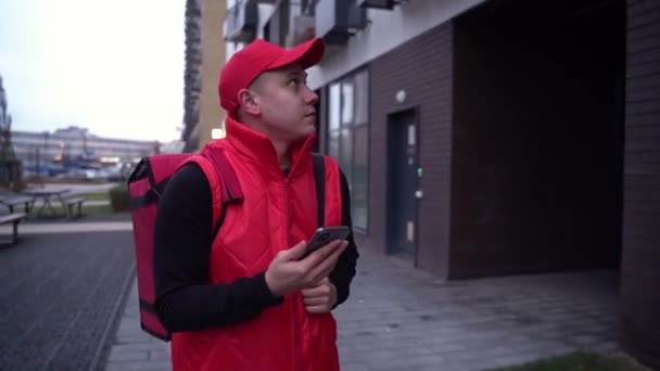 Leverans kille bär röd uniform när du går längs moderna byggnader ner city street med termisk väska och kontrollera kundens adress på sin telefon. Koncept för livsmedelsleverans — Stockvideo