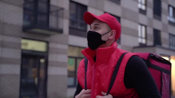 Pošťák v červené uniformě a ochranné masky při procházce po moderních budovách po ulici s termální taškou. Zpomalení střední střely — Stock video
