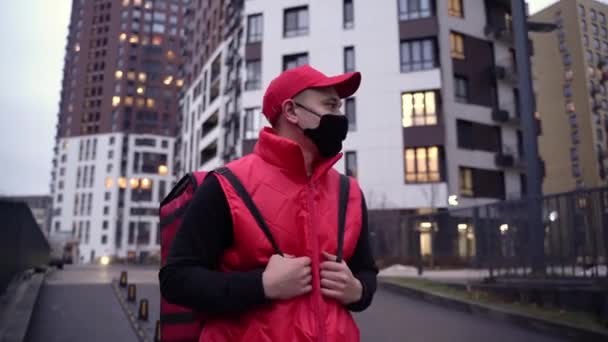 Курьер в красной униформе, прогуливаясь по современным зданиям по городской улице с термальной сумкой. Закрыть вслед за Шотом. — стоковое видео