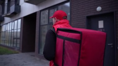 Şehir caddesinde termal çantalı, modern binalarda yürürken kırmızı üniformalı bir teslimatçı. Yakın çekim takip.