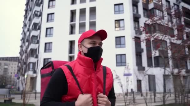 Repartidor con uniforme rojo y máscara negra mientras camina por edificios modernos calle abajo con bolsa térmica. Concepto de entrega — Vídeos de Stock