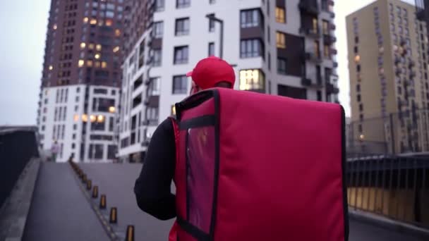 El repartidor vistiendo uniforme rojo mientras camina por edificios modernos calle abajo con bolsa térmica. De cerca seguir Shot — Vídeos de Stock