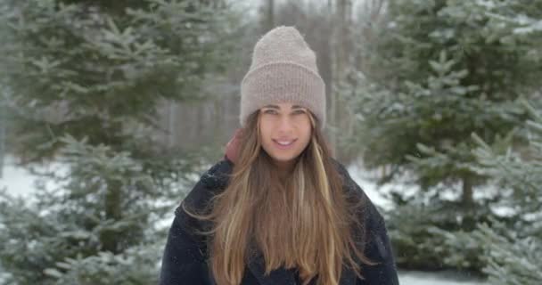 年轻美丽的女人站在冬季雪地的森林里看着相机。外头漂亮的帽子结霜寒冷的雪.靠近点肖像。慢动作 — 图库视频影像