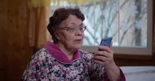 Η γιαγιά με το τηλέφωνο στα χέρια της. Μια ηλικιωμένη γιαγιά μιλάει μέσω video link με τους συγγενείς της σε κοντινό πλάνο Η ευτυχισμένη ηλικιωμένη γυναίκα μιλάει μέσω video link με την οικογένεια λόγω της κοινωνικής της απόστασης — Αρχείο Βίντεο
