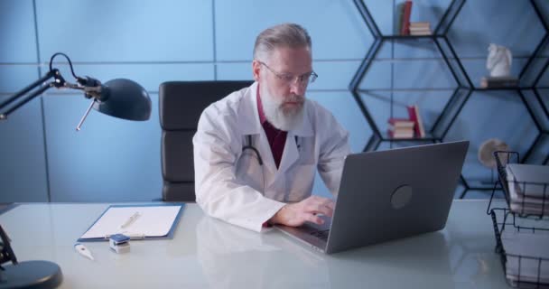 Médecin plus âgé dans le bureau travaillant sur un ordinateur portable et regardant la caméra. Portrait intérieur sérieux confiant thérapeute de famille en robe blanche au travail. — Video