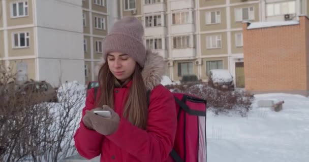 Entrega mujer que lleva con el uniforme rojo del teléfono inteligente mientras camina a lo largo de los edificios calle abajo de la ciudad con bolsa aislante — Vídeos de Stock