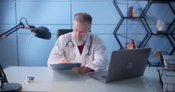 Médico de familia de mediana edad serio con una barba está trabajando en una clínica de salud. Médico sénior exitoso en bata blanca de laboratorio mira a la cámara en la oficina del hospital. — Vídeo de stock