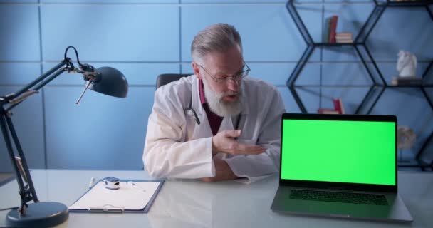 Семейный врач с бородой указывает на ноутбук с зеленым экраном в поликлинике. Профессиональный врач рядом с хромаки ноутбук в своем офисе — стоковое видео