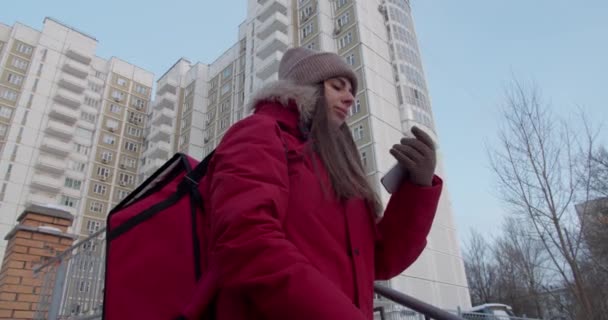 Een bezorger in een rood uniform kijkt naar haar telefoon en zoekt een afleveradres met een geïsoleerde tas. — Stockvideo