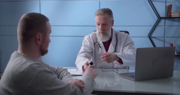 Professional serious focused middle aged hoary physician in medical coat sitting at table, consulting male patient about illness or surgery. Doctor used pulse oximeter on a patient during an — Stock Video
