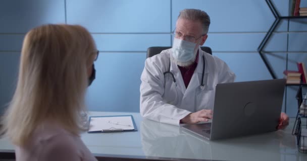 Professional serious focused middle aged hoary physician in medical mask and coat sitting at table, consulting female patient about illness or surgery. Woman listening to old doctor at checkup meeting — Stock Video