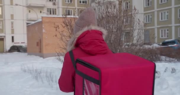 Suivez la photo de la femme de livraison portant un uniforme rouge smartphone tout en marchant le long des bâtiments dans la rue de la ville avec un sac isolé. Vue arrière — Video