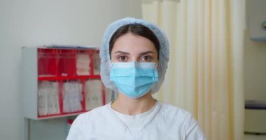 Tıbbi üniformalı kadın hemşirenin cerrahi maskesini çıkarırken, kameraya bakarken ve ağır çekimde gülümserken bir portre resmi. Hastanedeki maske takan güzel beyaz kadın..