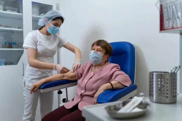 Perawat yang mengenakan sarung tangan pelindung dan mask pelindung memberikan suntikan Vaksin kepada pasien wanita tua di klinik medis sungguhan selama pandemi. Dokter wanita memakai masker medis dan sarung tangan. Stok Gambar Bebas Royalti