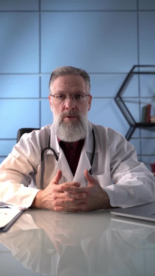 Vertikales Video. Professioneller männlicher Arzt mit Bart in weißer medizinischer Uniform, der in die Kamera schaut, führt eine Videokonferenz oder zeichnet ein Webinar zur Ausbildung im Gesundheitswesen auf. — Stockvideo