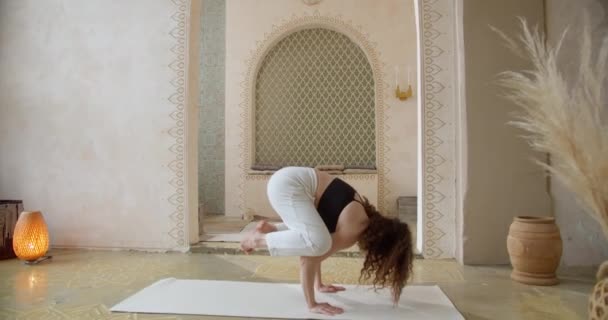 Спортивная кудрявая женщина занимается упражнениями по йоге. Стройная девушка занимается йогой в яркой, атмосферной комнате для йоги утром — стоковое видео
