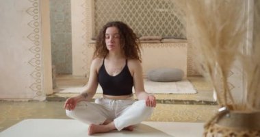 Spor giysili rahat kıvırcık saçlı genç kadın sabah atmosferik yoga odasında gözleri kapalı yoga minderinin üzerinde meditasyon yapıyor. Sportif kız elleriyle meditasyondan zevk alıyor.