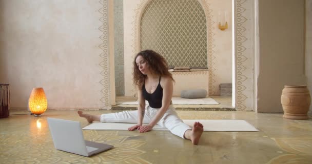 Joven mujer de pelo rizado estirando el cuerpo viendo la lección en línea sentado en la esterilla de yoga. Aprendizaje y estudio en línea, estilo de vida saludable activo, entrenamiento deportivo femenino, bienestar y ejercicio en interiores — Vídeos de Stock