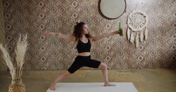 Yoga de la cerveza. Mujer joven de pelo rizado haciendo ejercicio de estiramiento de yoga con una botella de cerveza. Deportiva chica delgada haciendo yoga del vino en una sala de yoga luminosa y atmosférica por la mañana — Vídeos de Stock